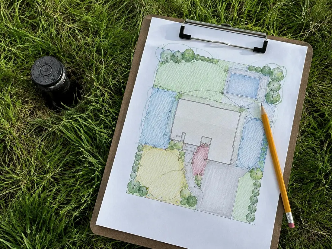 A hand-drawn irrigation zone plan on a clipboard resting on the grass beside a sprinkler head